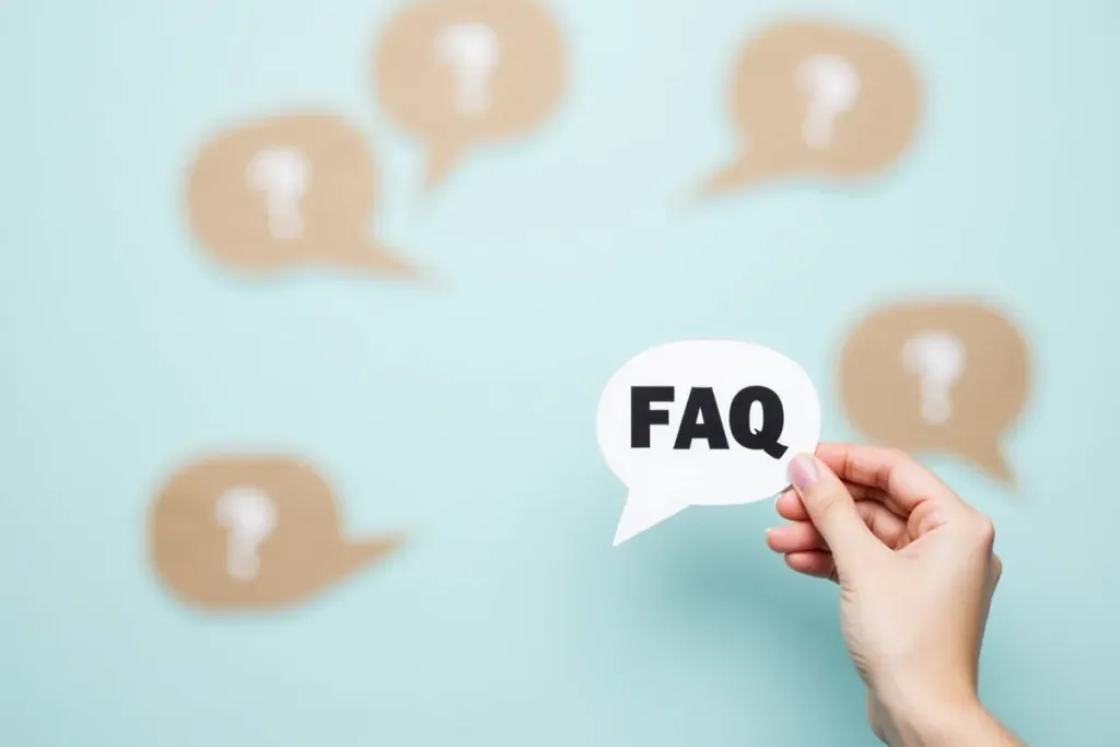 Eine Hand hält eine Sprechblase mit der Aufschrift 'FAQ', während im Hintergrund mehrere Sprechblasen mit Fragezeichen zu sehen sind. Das Bild symbolisiert häufig gestellte Fragen und Antworten.