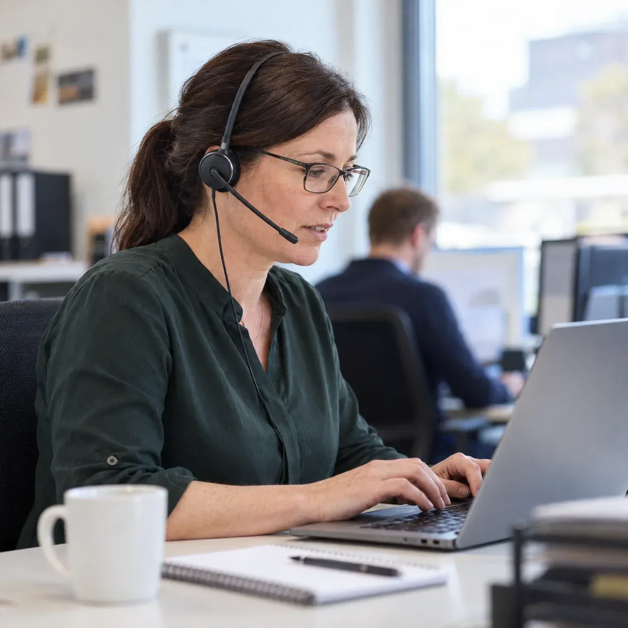 Lächelnde Frau mit Kopfhörern, sitzt an einem Schreibtisch mit Laptop in einem hellen Büro, Sekretariatsservice.