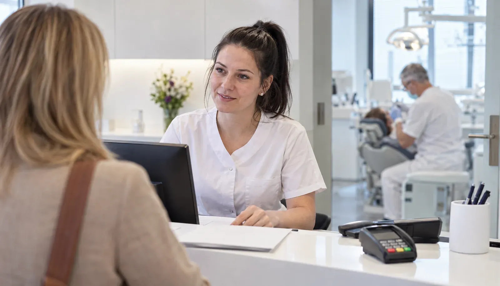 Zahnmedizinische Fachangestellte begrüßt eine Patientin an der Anmeldung, im Hintergrund der Zahnarzt